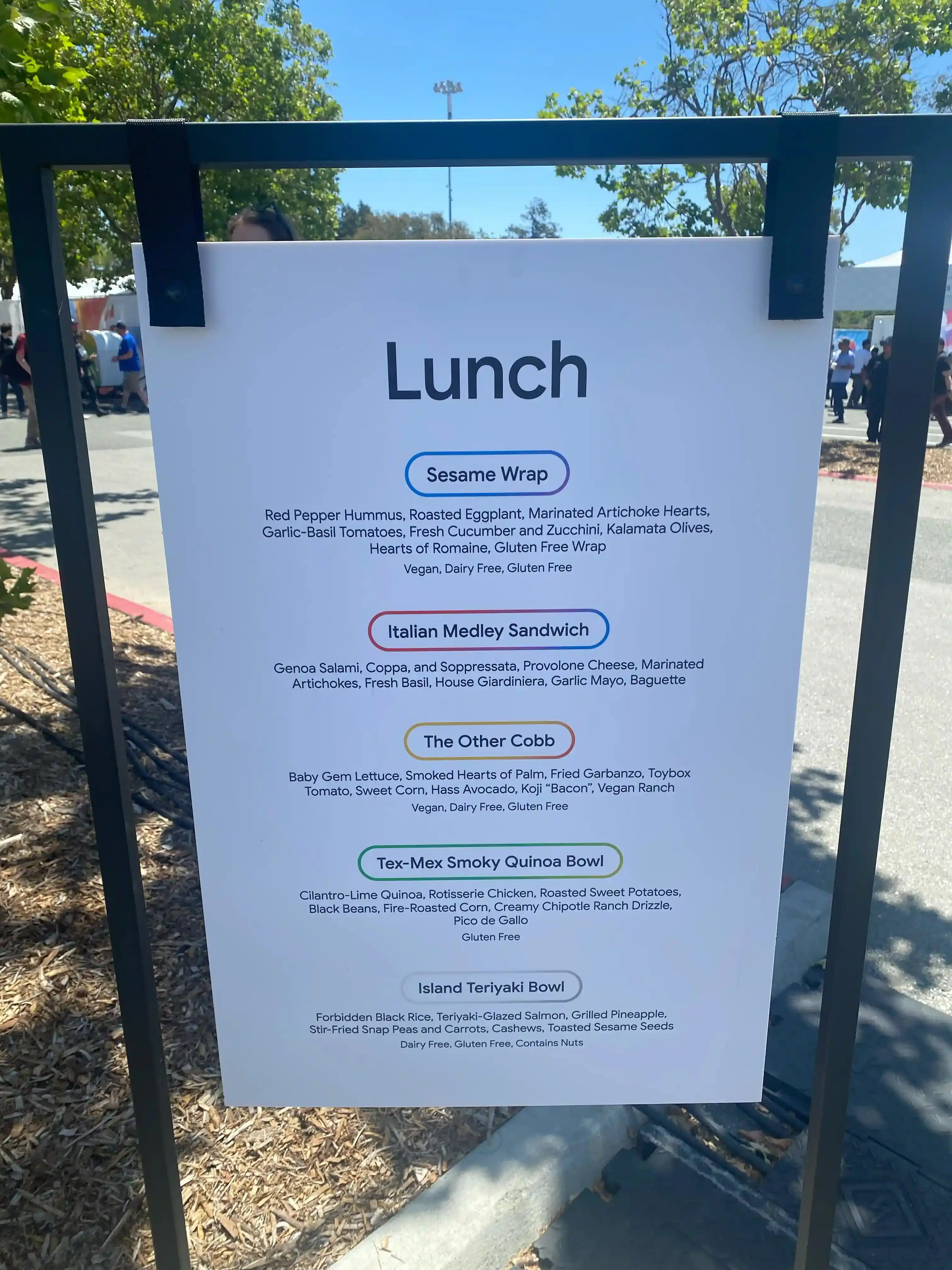A sign listing lunch menus. There are five items: sesami wrap, italian medley sandwich, the other cobb, tex-mex smoky quinoa bowl, and island teriyaki bowl. Detailed ingredients and whether each is vegan or gluten-free are also listed.